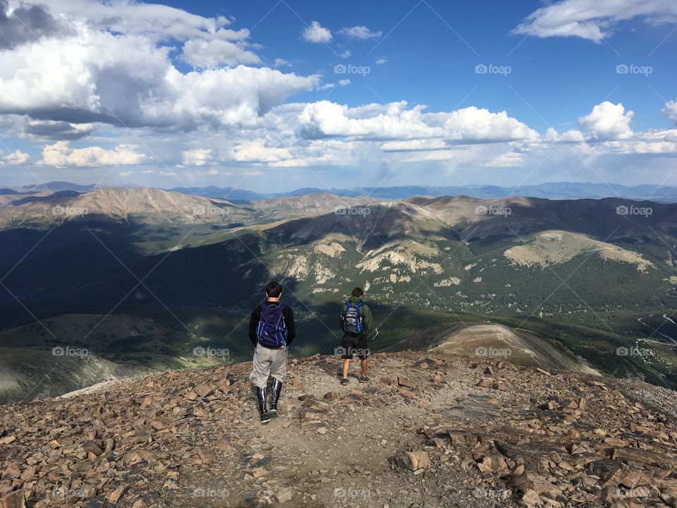 Two men hiking along a rocky path on Quandary Peak in Colorado surrounded by mountains, blue sky, and clouds