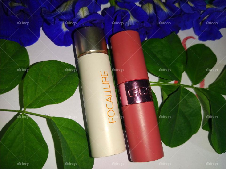 Lipsticks in the middle of green leaves and fresh blue flowers against a white background.