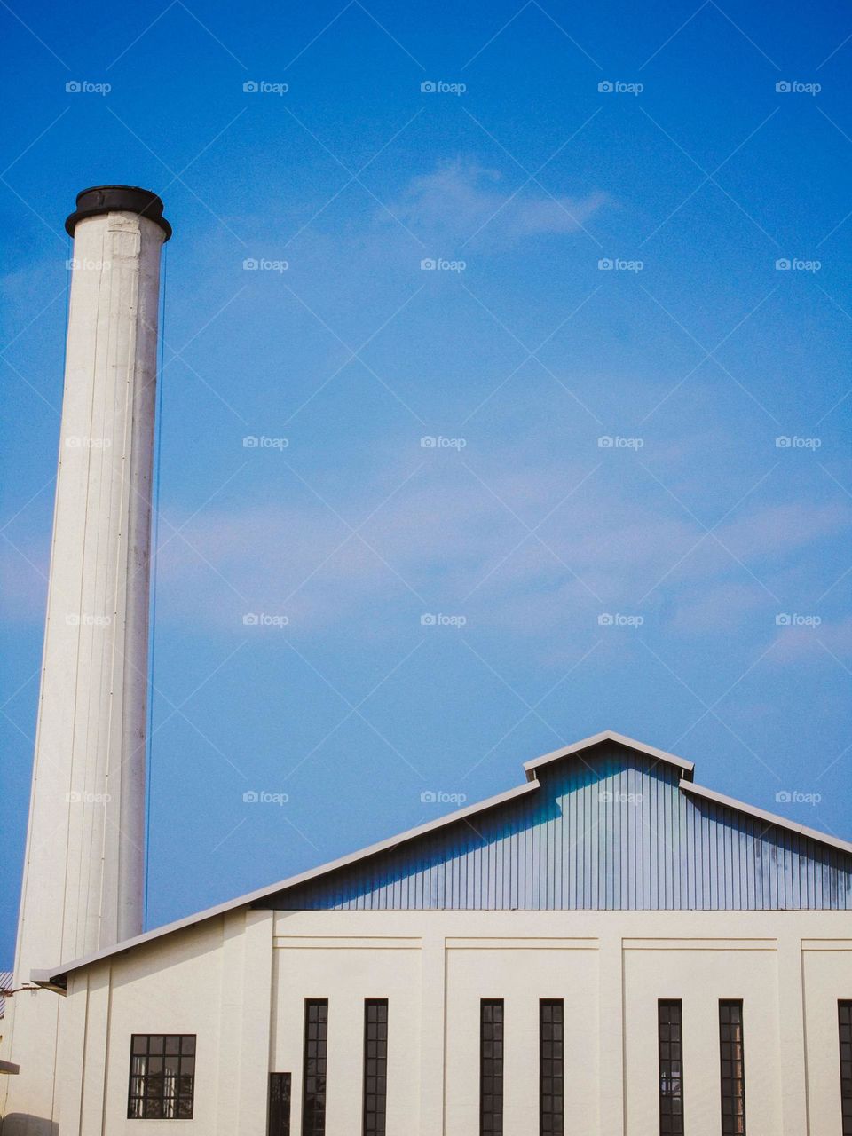 Close-up view of a white building with a blue roof and tall chimneys in the foreground, with a blue sky and some clouds in the background