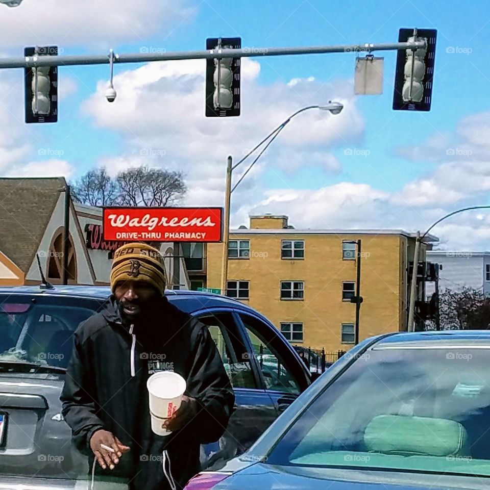 Panhandling in City traffic, cars stopped at lights overhead. He's carrying a cup for drivers to put money into. Knit hat on mans head, cold weather out.