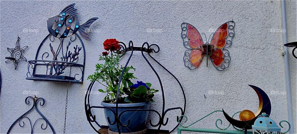 These ornamental cast iron hangers hold potted plants and flowers in full bloom on the terrace of a beautiful house in rural “Bavaria”, Germany. Handcrafted oversized butterflies complete this beautiful springtime motif. 2024. Hypnotic Productions