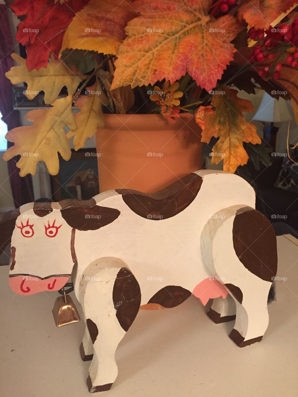 Wooden painted  whimsical black and white cow sitting in front of a clay tall container of fall flowers. 