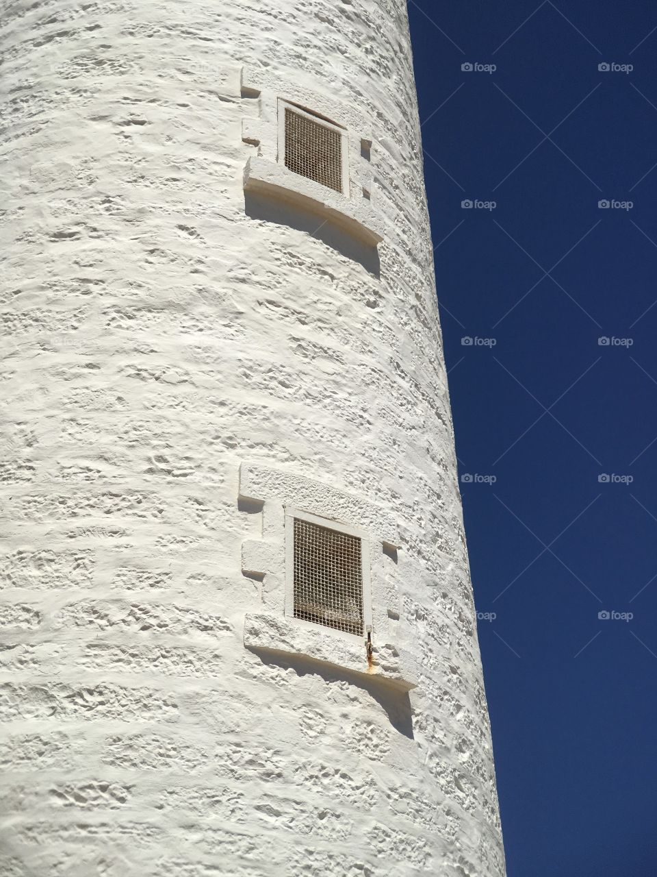 Minimalist image old stone lighthouse side wall of windows looking out toward the ocean on clear vivid blue day 