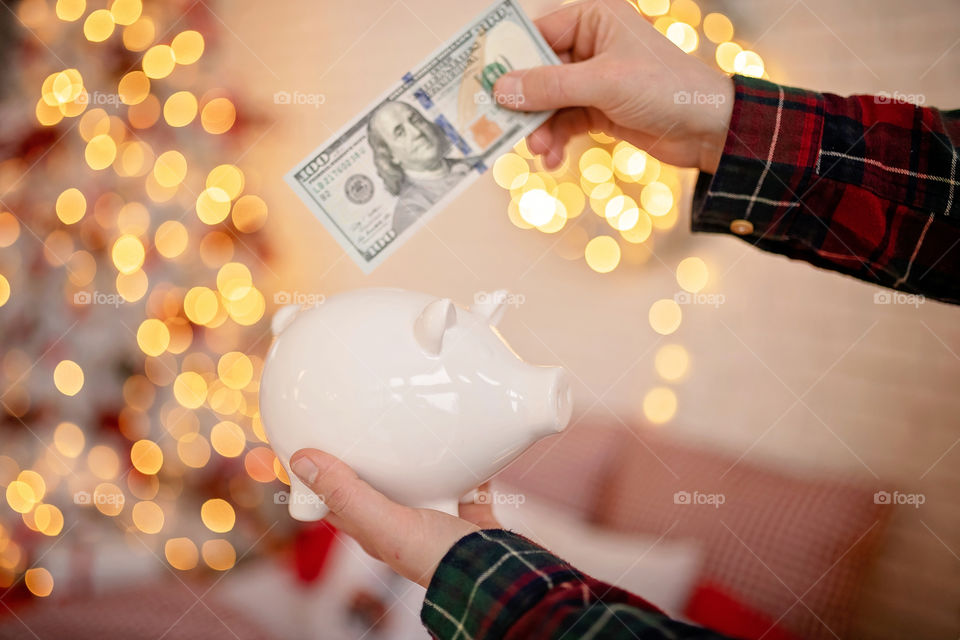 piggy bank with 100 dollars banknote in festive New Year atmosphere of scenery.  Magical bokeh with Christmas tree and bright lights.