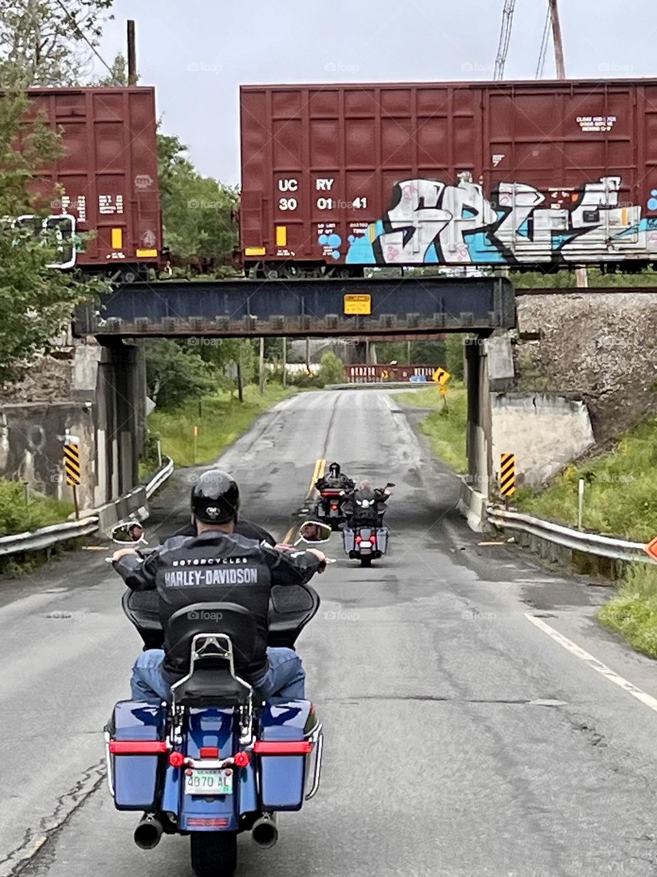 Motorcycle riders passing underneath a train trestle with train cars covered in graffiti 