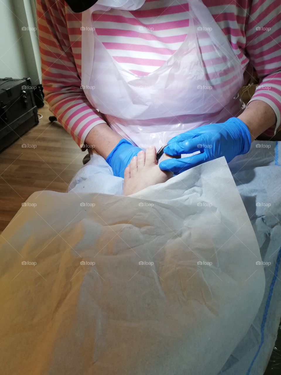 A woman in a pink white striped shirt making pedicure, wearing a plastic apron and blue gloves, holding a foot of a client between her hands, the rest of the body is protected with a white cover.