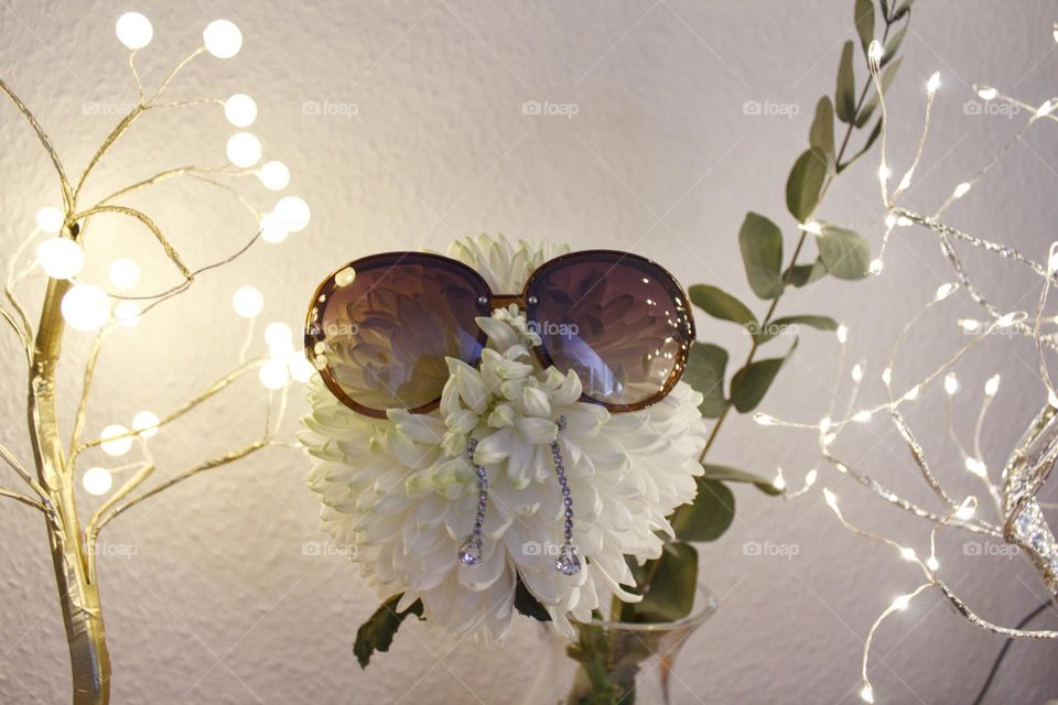 White chrysanthemum with sunglasses and earrings in front of a white wall with eucalyptus and light trees.