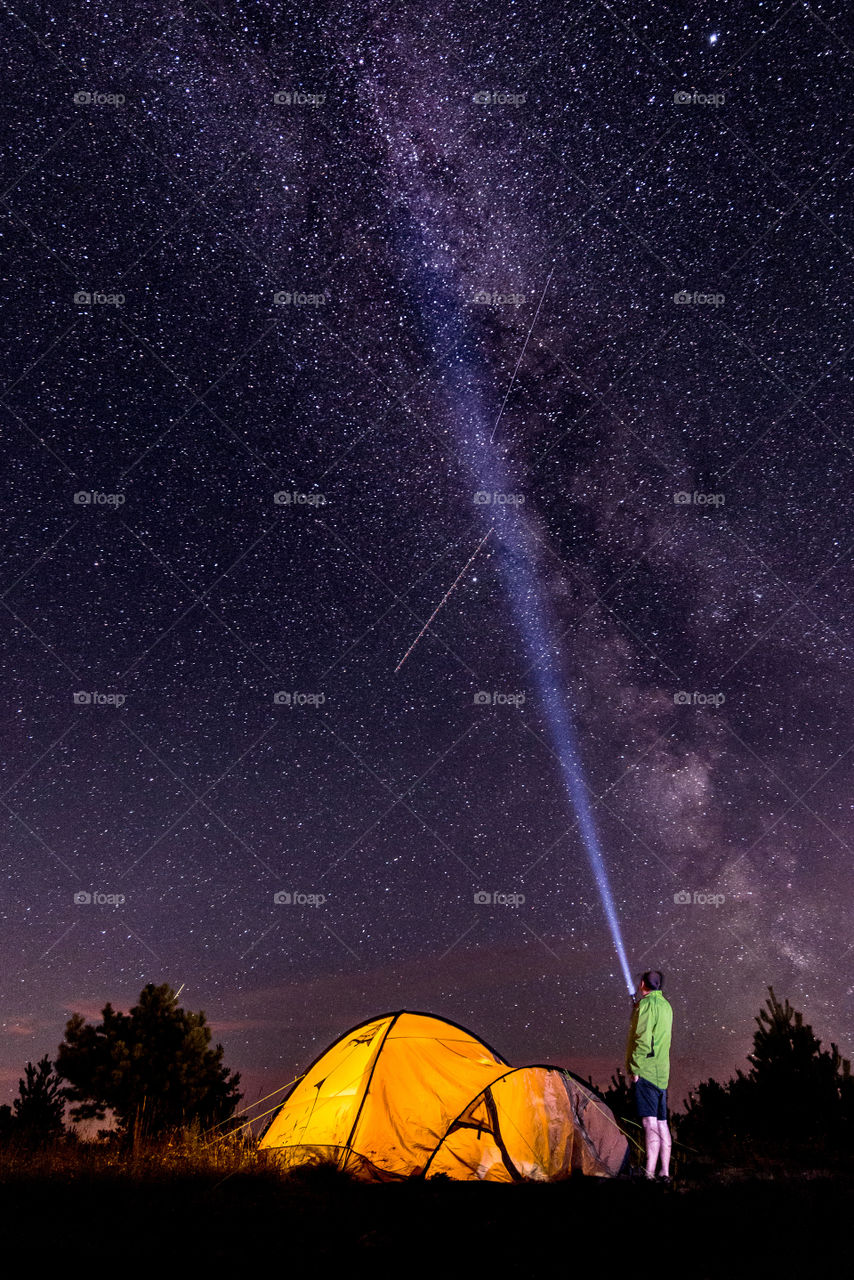 At night a man stands in front of an iluminated tent and sends a ray of light into the starry sky, milky way
