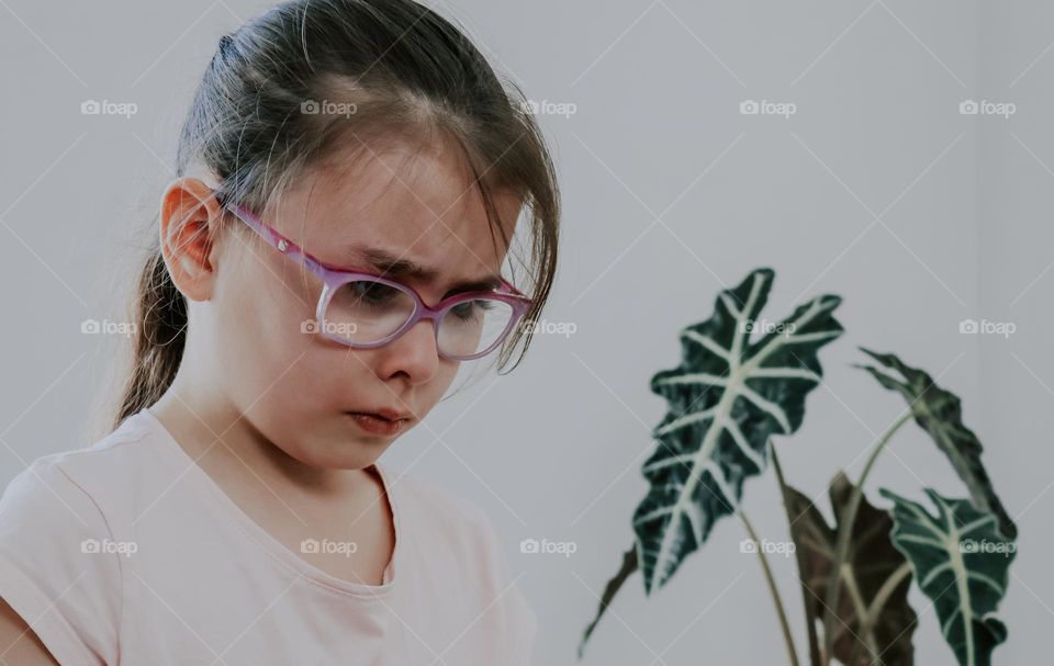 Portrait of a beautiful caucasian brunette girl in glasses with a sad and offended emotion to tears on her face, stands in a room opposite a window with a flower, close-up side view. Sad people concept.