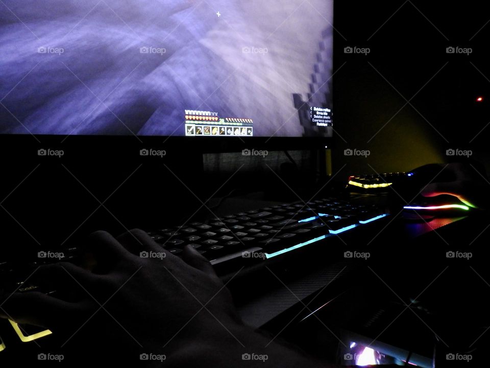 Teenager boy playing Minecraft on the gaming computer seen from his left side and close-up of his hand playing in a dark room.