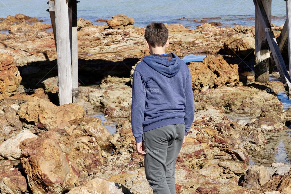 12 year old boy wearing hoodie jacket walking across rocks toward ocean Jetty wharf dock 