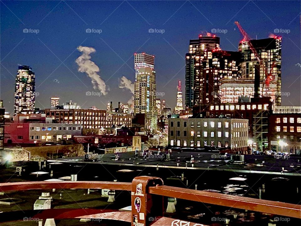 A nighttime shot of the Long Island City, Queens, NY skyline as seen from the Pulaski Bridge at night with two chimneys releasing clearly visible smoke stacks into the sky. 2022. Hypnotic Productions