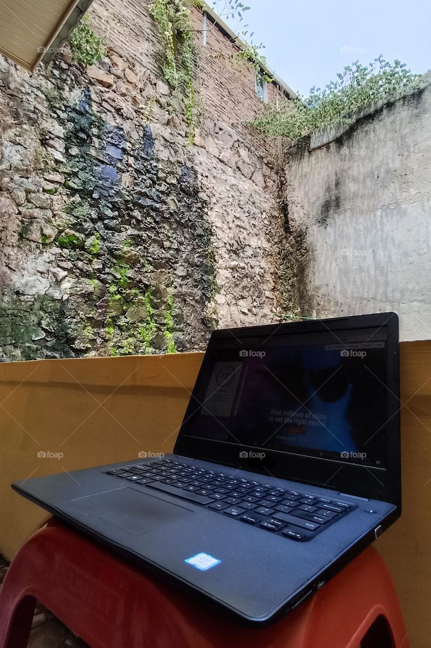 a laptop that is open and placed on a red plastic bench, against the background of a stone wall overgrown with moss and plants