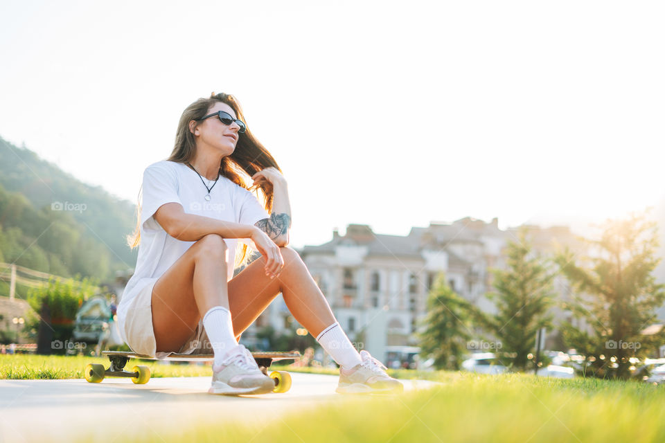 Slim young woman with long blonde hair in light sports clothes with longboard in outdoor skatepark at sunset