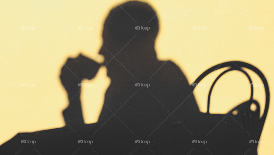 The grey shadow on a person drinking morning coffee is cast on onto a pale yellow wall