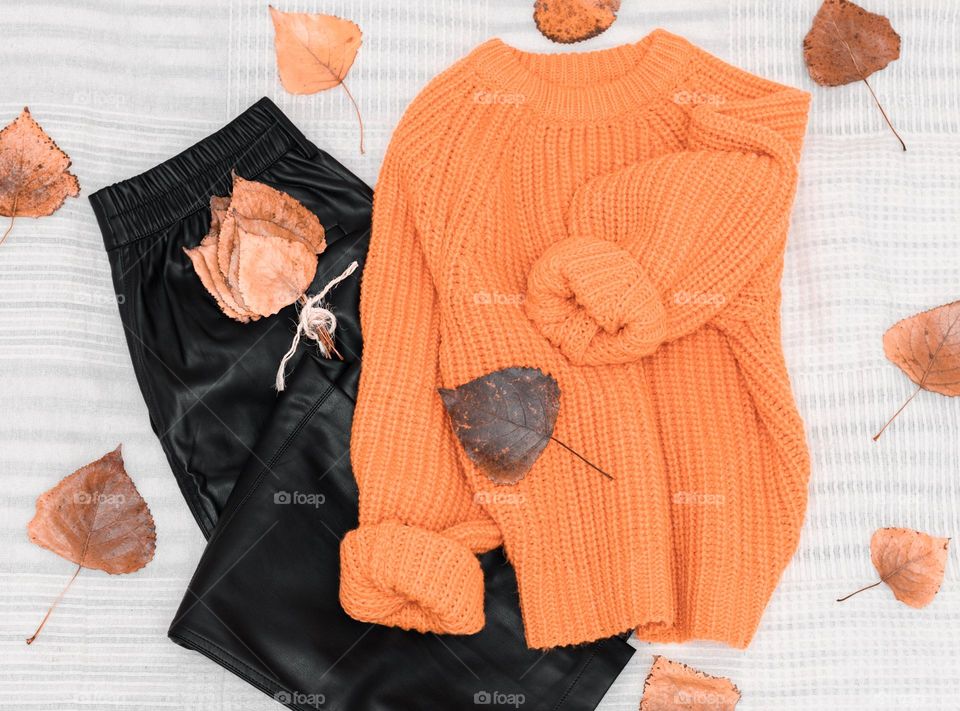 An orange knitted sweater and black leather pants with poplar leaves scattered around autumn lie on a bed with a white bedspread, flat lay close-up. The concept of fashionable demi-season clothes, autumn collection.