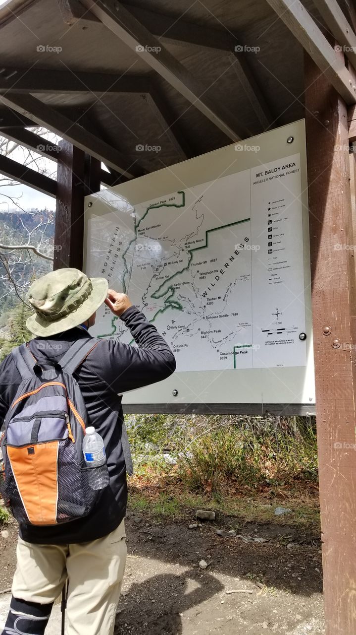 man wearing backpack and a hat pointing at a trail map for a mountain hiking during the winter, spring, summer, fall season