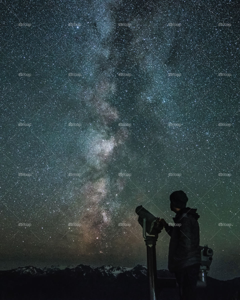 Silhouette of a person next to a telescope, as the Milky Way shines brightly in the dark night sky during late summer. 