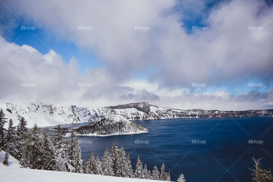The water at a snow covered Crater Lake National Park is glowing bright and bold blue beneath a partly cloudy sky. The pine trees as far as the eye can see are dusted with a pure white snowfall. 
