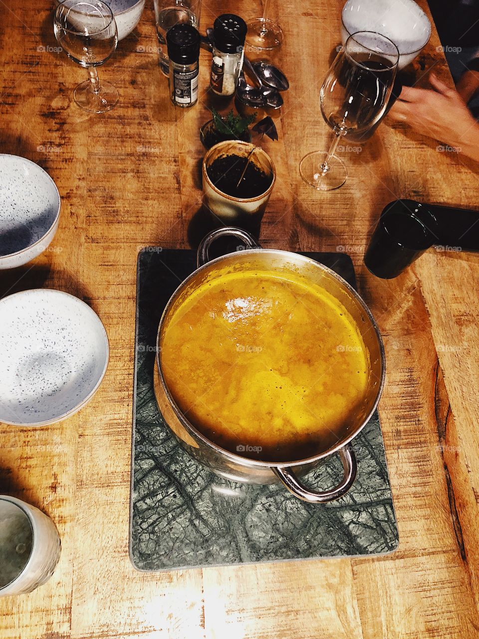 Family dinner time with one pot vegetable soup supper in stainless steel pan on green marble trivet and wooden table background. Some glassware and tableware visible and decorative small pot plant in centre