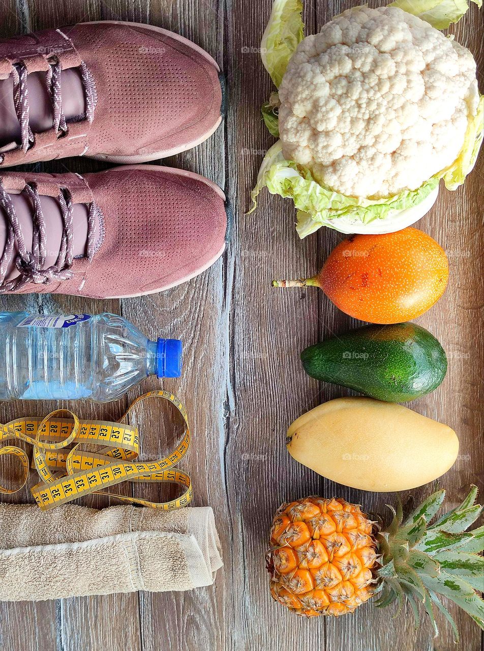 The wooden background is invisibly divided: on the one hand, fruits and vegetables, on the other hand, healthy lifestyle accessories. Cauliflower, granadilla, avocado, mango and pineapple. Sneakers, measuring tape, towel, bottle of water