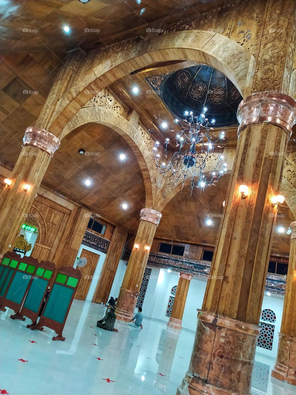 The interior of a magnificent mosque, carved wooden pillars, luxurious chandeliers, white tiled floors and several people praying.