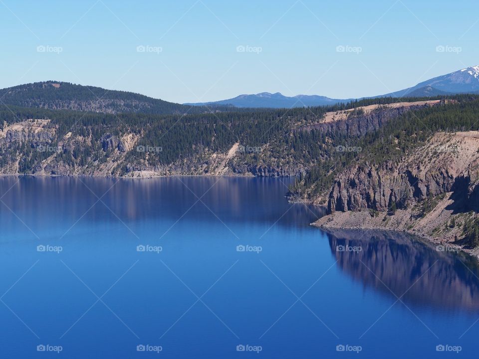 The rich blue waters of the deep Crater Lake in Southern Oregon with fir trees on the jagged rim on a beautiful sunny summer morning with clear blue skies. 