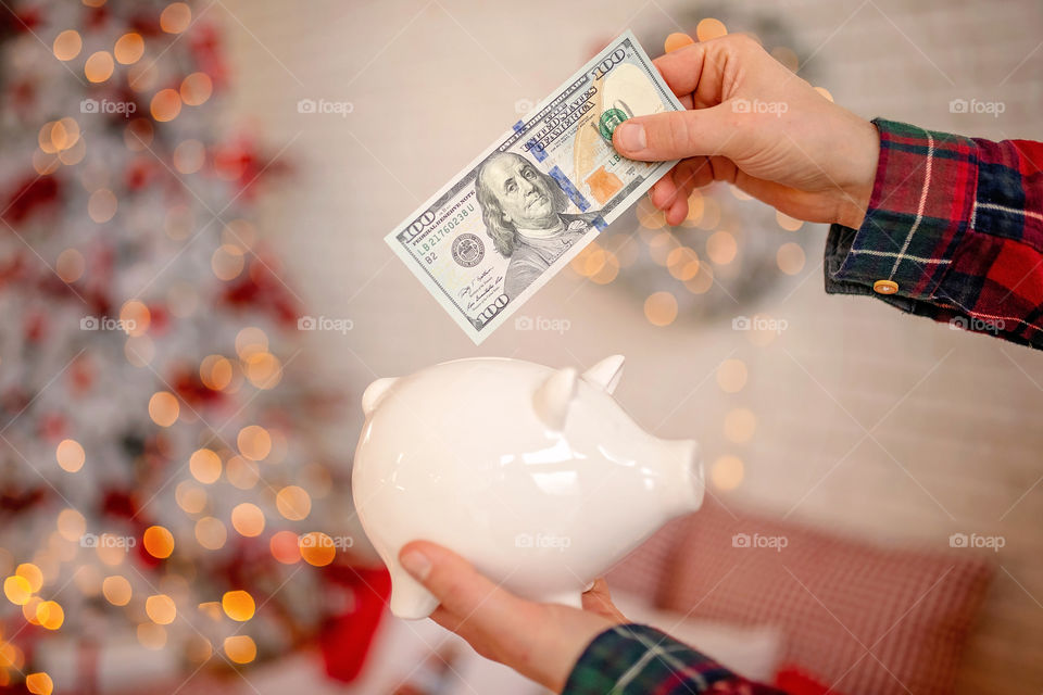 piggy bank with 100 dollars banknote in festive New Year atmosphere of scenery.  Magical bokeh with Christmas tree and bright lights.