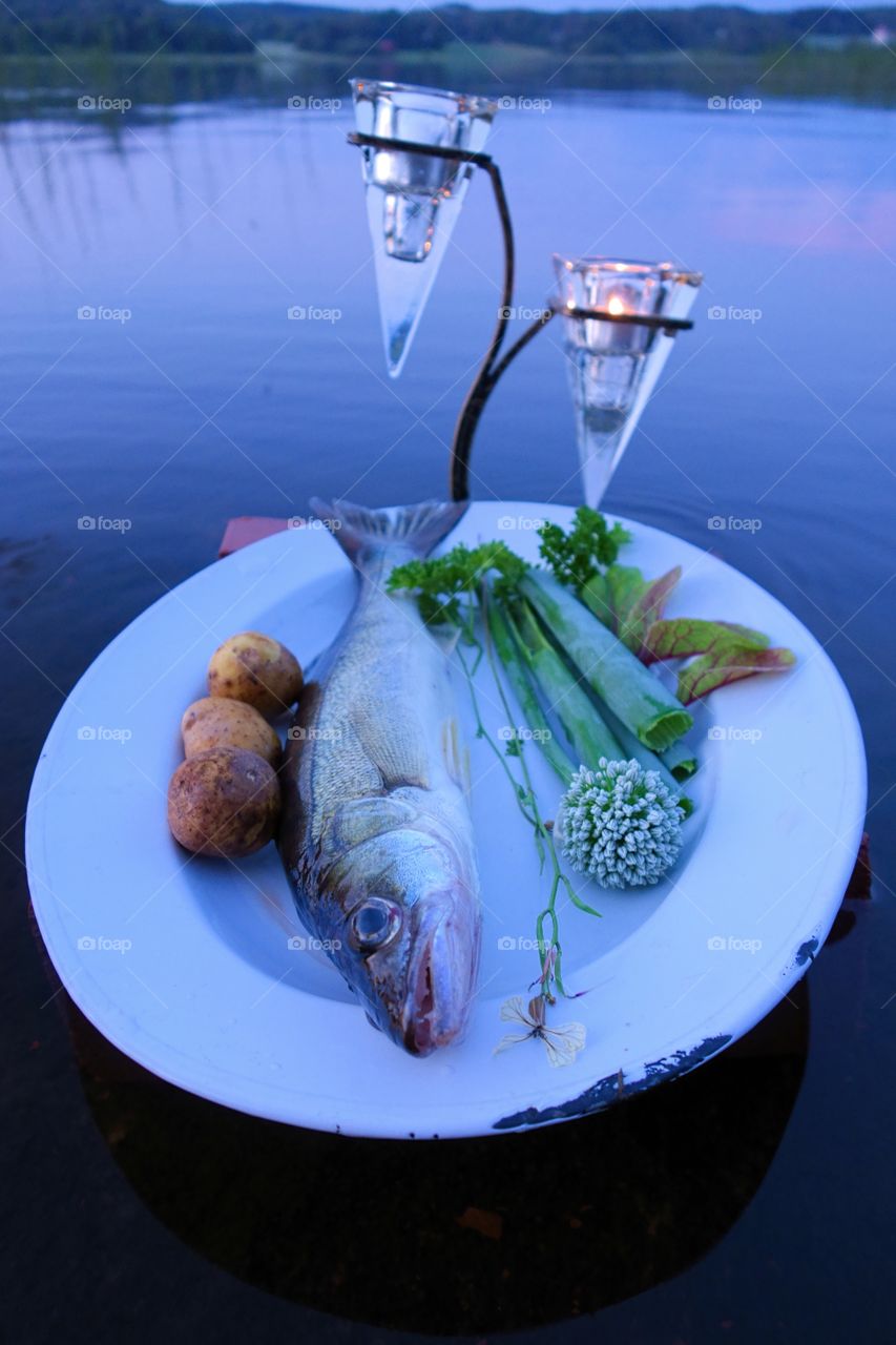 
Fresh fish catch on a plate with vegetables above water by the lake in summer evening in Finland.. Fresh fish catch (pike perch) on plate served with fresh vegetables in outdoors setting on a lake with candles as a decoration.