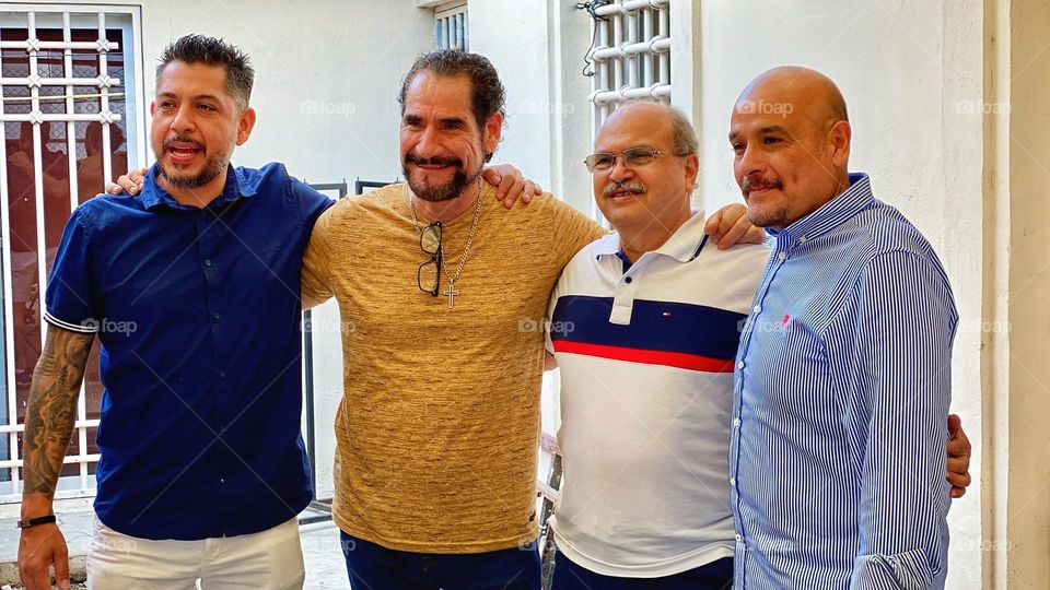 Group of men smiling and hugged between them, posing for a portrait picture taken from another angle.