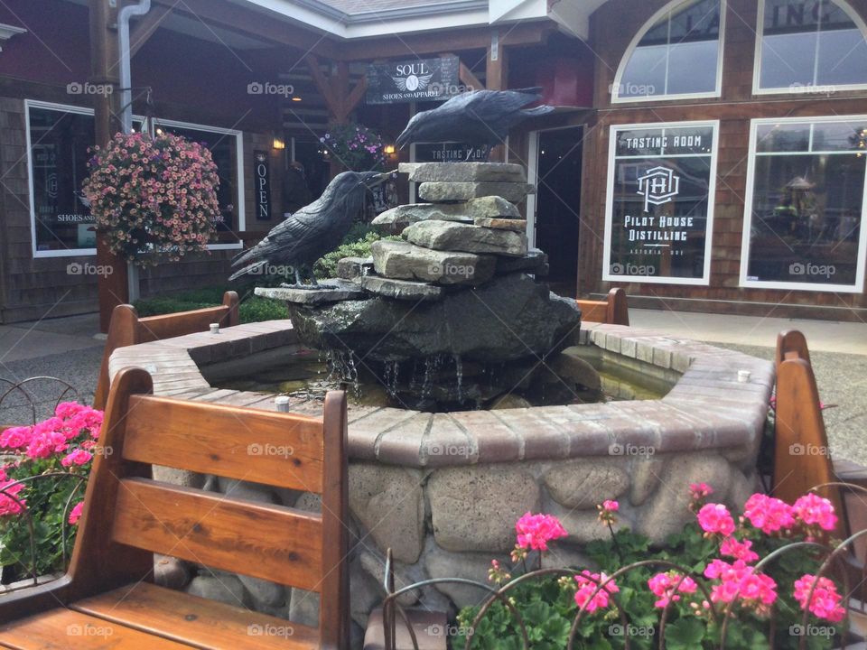 A Water Fountain with two Sculptures of Crows in Cannon Beach, surrounded by planters with pink flowers 