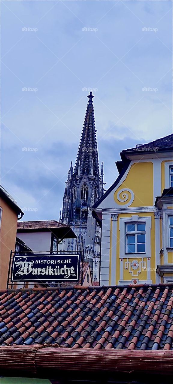 This is the old city of “Regensburg” by the river “Danube”. We see the gothic steeple of the cathedral and the sign in black letters invites to the “Historische Wurstküche”, the 1200 year old “Historical Sausage Kitchen”. 2024. Hypnotic Productions
