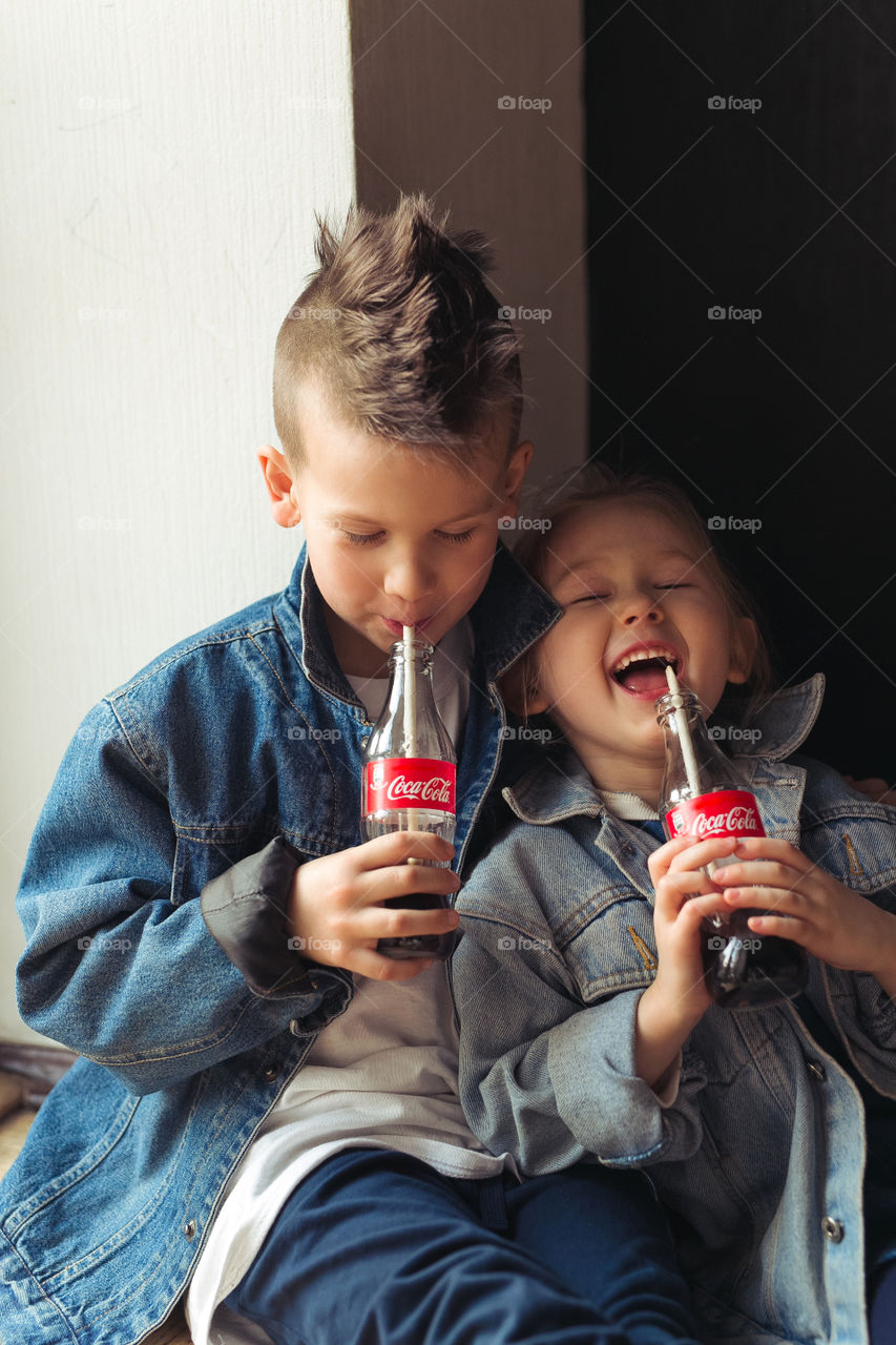 a boy and a girl of seven and five years old, friends, drinking Coca Cola, laughing, having fun, wearing denim clothes. Emotionally. Lifestyle photo. Happy kids.