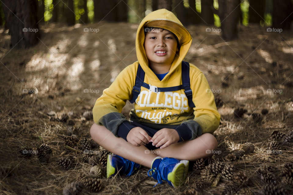 Seasonal outdoor portrait of a young happy Eurasian kid hiking in a pine forest wood.Natural setting, the boy is wearing blue sneakers, yellow sweater, hood and cap, smiling while looking at the camera, seating on the floor surrounded by pinecones