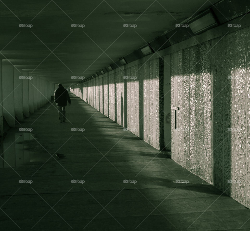 Long shadows are cast along an underground walkway, one man walking can be seen in the distance in greyscale with hints of green