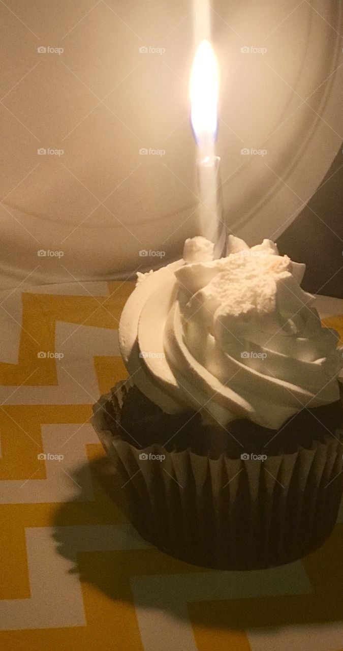 Vanilla frosting on a yummy chocolate cupcake with a bright and shiny glowing candle on a yellow checkered paper Birthday plate 