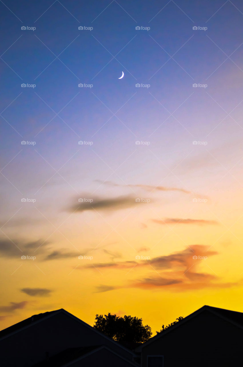 Small crescent moon in the sky during sunset above the rooftops of two houses