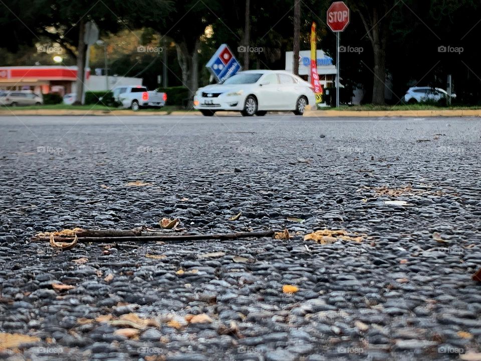 Commuting: vehicles busy going on their own ways and direction where they need to be and go down the road and in a parking lot entrance seeing businesses, trees and stop signs.