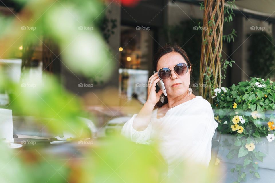 Beautiful brunette middle aged woman in sunglasses in white clothes talking on mobile phone in hand with coffee at terrace of the summer cafe