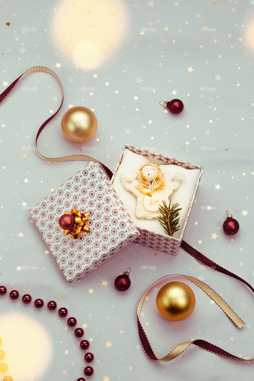 Wrapping Christmas gift into box. Decorating using red ribbon, red Christmas balls and pine twigs. Blurred Christmas lights. Flat top-down composition. Figure of angel made of earthen with love inscription as Christmas decoration