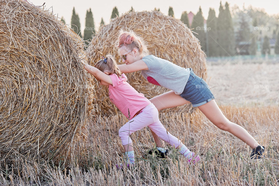 Sisters, teenage girl and her younger sister playing together with hay bale outdoors in the field in the countryside. Candid people, real moments, authentic situations