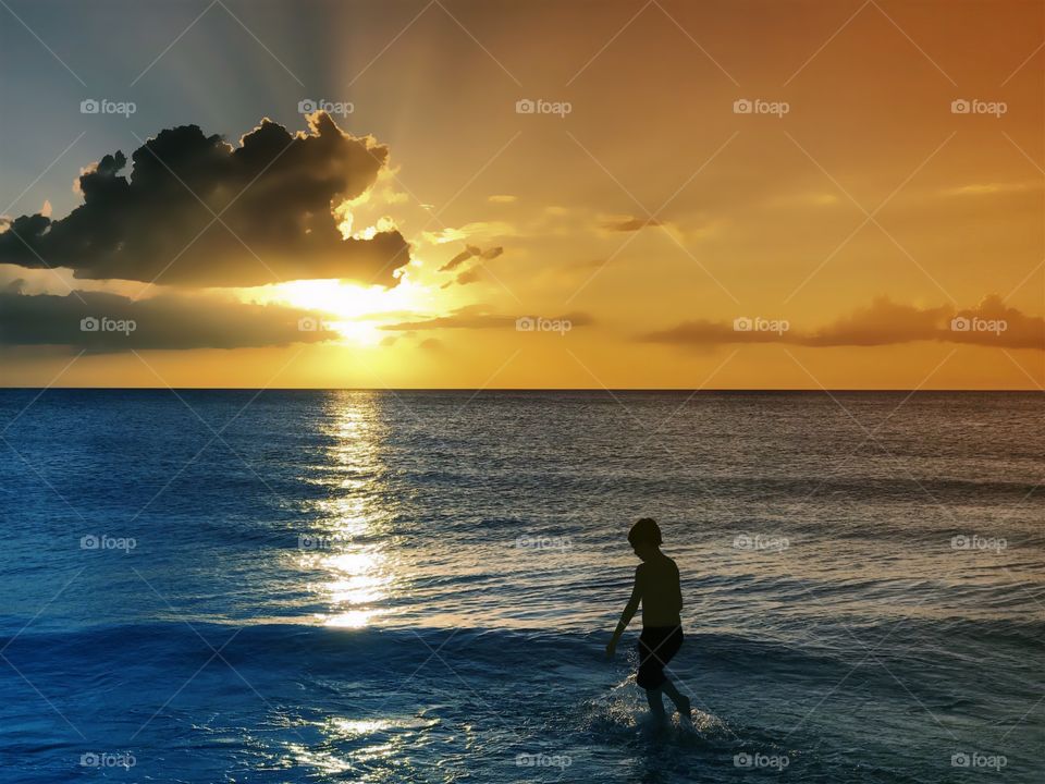 Small boy wading through the ocean under a spectacular golden sunset.