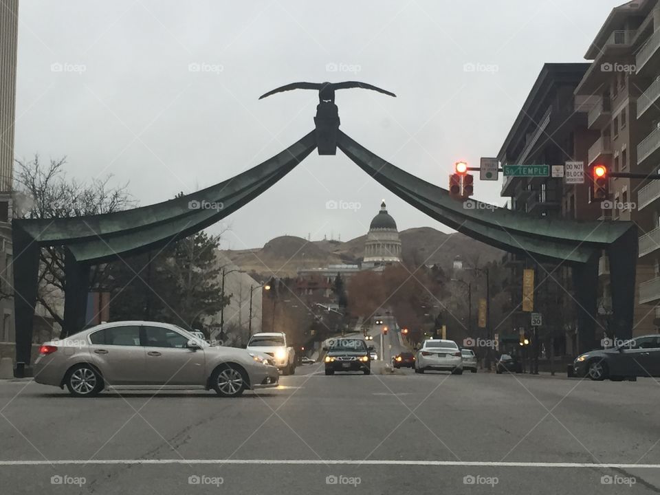 A monument in downtown Salt Lake City, Utah during winter. An arch with an eagle hangs over a busy street with the Capital building visible in the background.