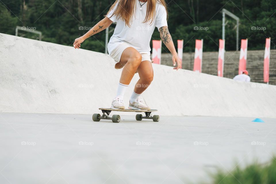 Slim young woman with long blonde hair in light sports clothes on longboard in outdoor skatepark at sunset time