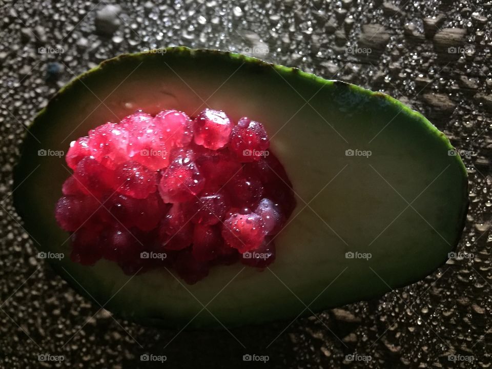 Half a wet green avocado with pomegranate seeds in the hollowed-out center. The avocado is sittting on a wet wood table and is side lit to highlight the drops on the table and show the translucency of the pomegranate seeds. 
