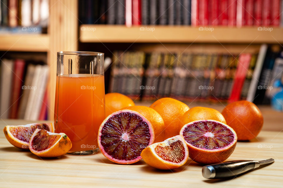 glass of juice, knife and cut red oranges on a light wooden table. Blurred book shelves in the background.