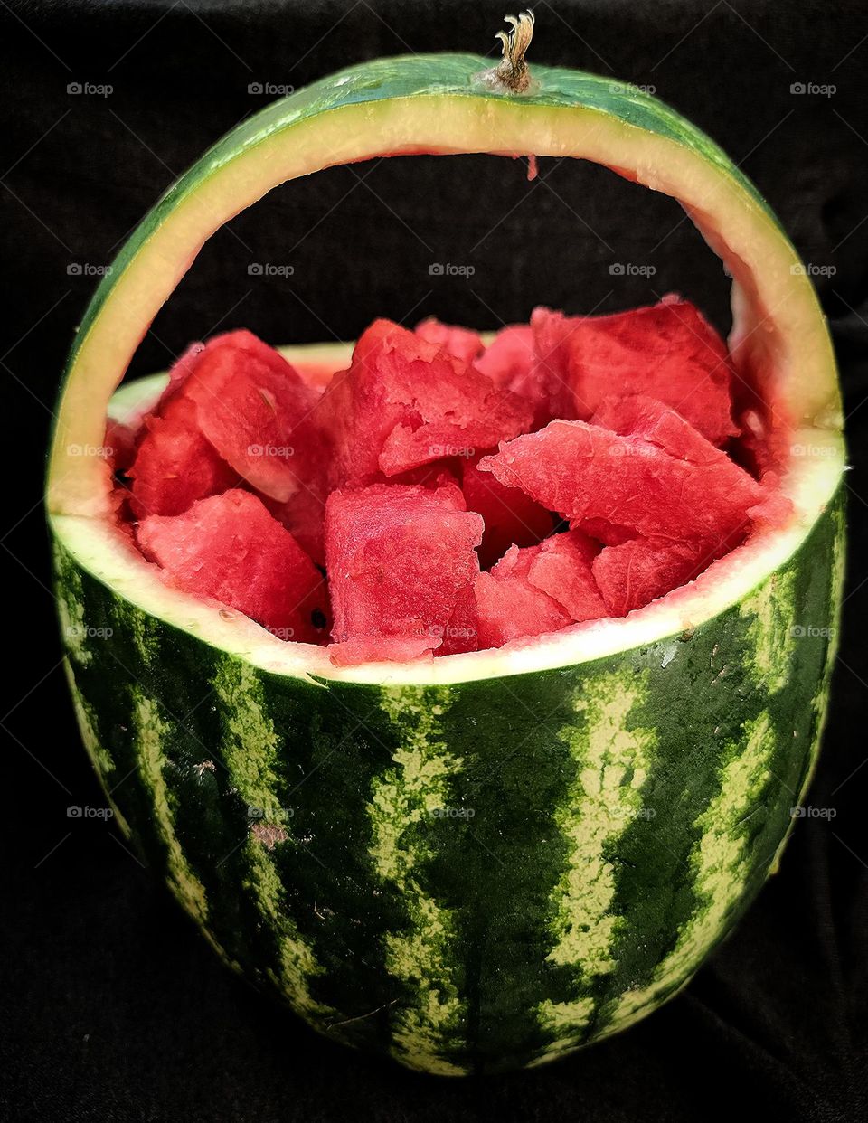 Summer treats.  A green watermelon rind with black stripes is cut into the shape of a vase with a handle.  Inside are juicy slices of red watermelon.  Black background