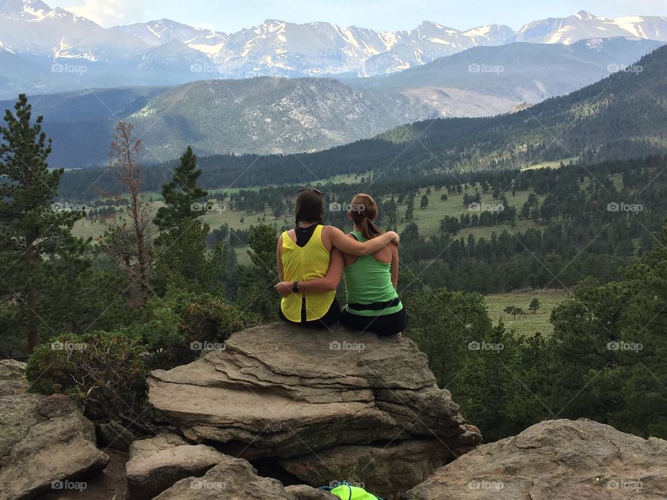 Friends forever. Two young women arm in arm, looking at the mountains and green valleys and sitting on a cliff. 