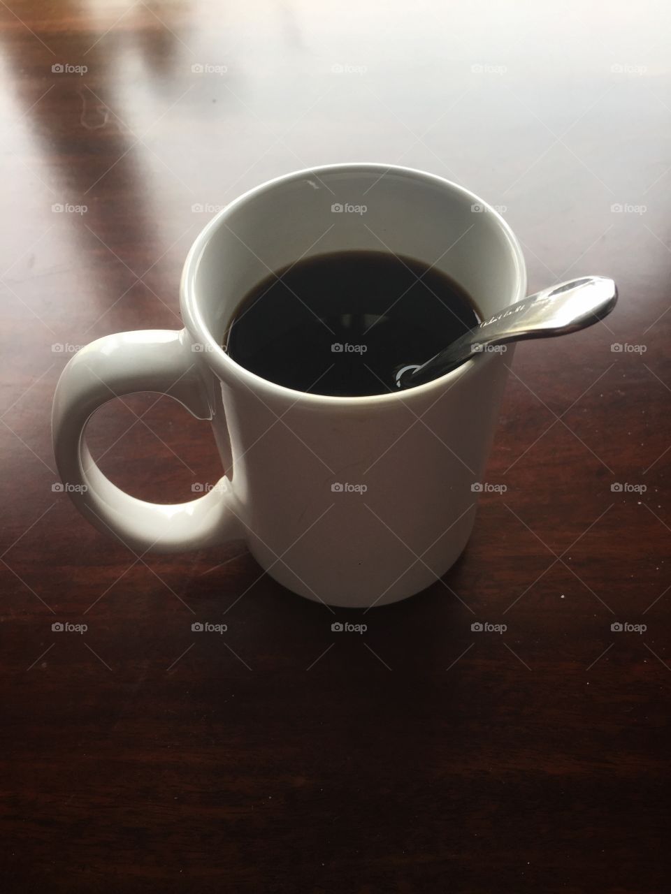 A cup of black coffee as seen from the side and above. The mug is white and there is a solver spoon in the coffee.