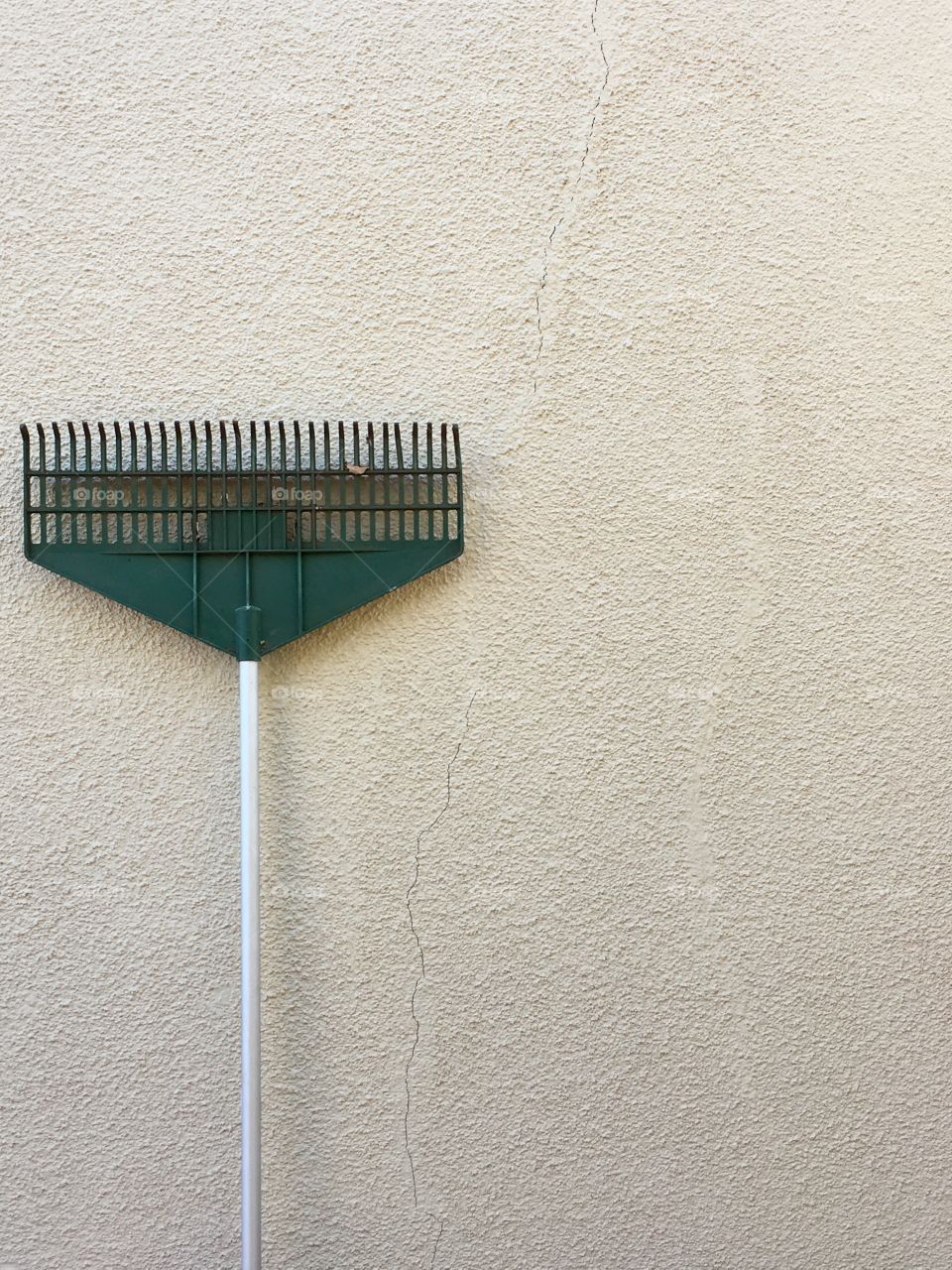 Garden rake leaning up against old exterior stucco wall which has a crack running through it. Rake to left of image, negative space minimalist image 